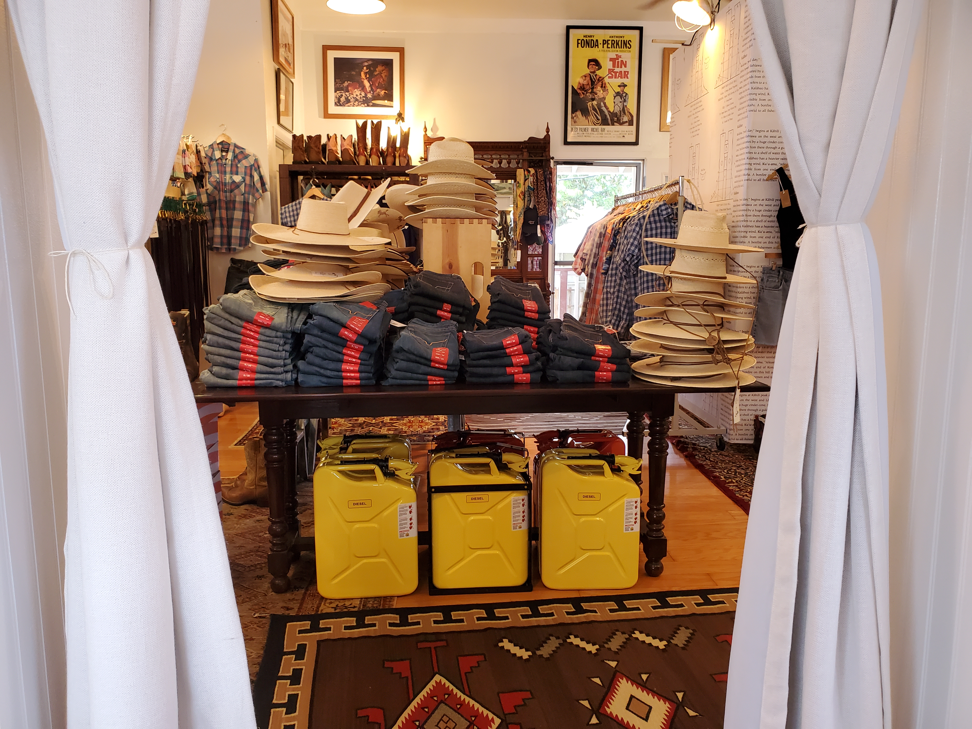 Photo of store after entering through front doors and passing through foyer. Soft warm lighting, large room, tall ceilings. Interior shows racks, tables and shelves of products. There are five bright yellow metal 5 gallon NATO diesel fuel cans and one bright red gasoline can lined up on the floor under a wooden table with stacks of many different styles and sizes of Kimes ranch blue denim jeans. Also arranged in stacks by style are Sunbody straw and grass hats arranged on the table at different heights. In the background are shelves of vintage cowboy boots. Next to the boots are hooks filled with Tory leather belts of different colors and sizes. Against the back wall is an antique Victorian entry hall bench with decorative finials, multiple mirrors and brass rails. Vintage and new bandanas and scarves decorate the bench. To the right is a free standing dressing room which is next to a rack of mens and womens Palaka shirts in long and short sleeves. Old posters on the walls of cowboys and new New Mexico style art framed. The wood floor is partially covered by hand woven wool rugs from Oaxaca and Persia. 