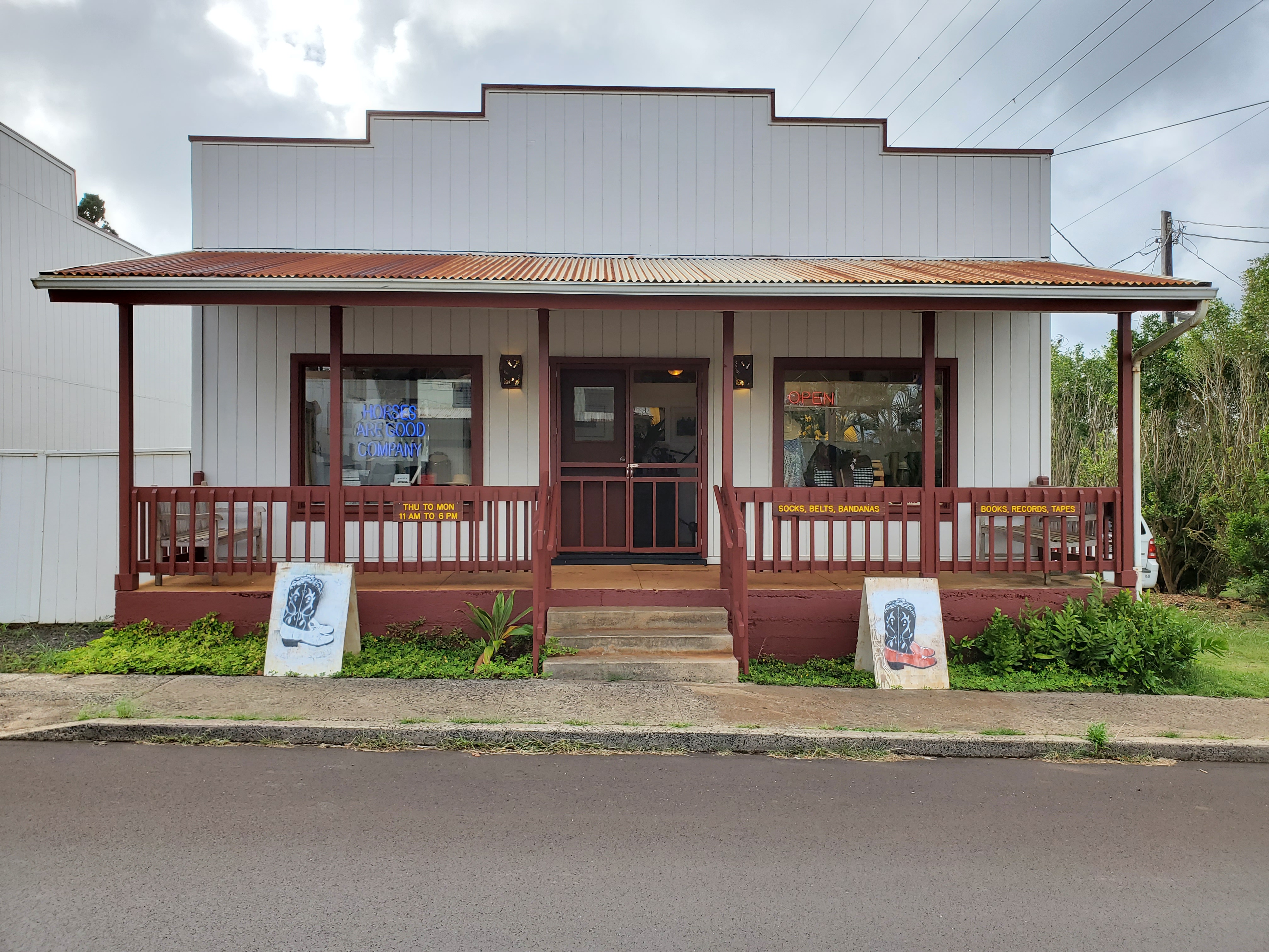 Photo of the Horses Are Good Company store front in Kalaheo, Kauai, Hawaii. Showing a four step walk up single story building with gray painted vertical wood siding and dark red trim. Glass French doors with additional screen doors at entrance. Exterior of commercial building has standing one on either side of the stairs, painted plywood sandwich boards decorated with multi-color stencils of cowboy boots with stitching. Storefront has four concrete steps leading up to covered porch with benches on either side. The railing and posts are all painted the same dark red. Through the large window on the left you can see a bright blue neon sign that says Horses Are Good Company. On the large window on the right there is a bright red neon sign that says Open. A vintage leather western style saddle is in the window below the Open sign. 